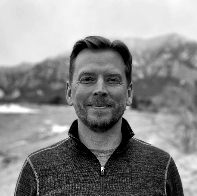 Black and white portrait Dan Getman wearing a zipped up jacked with mountain and water in the background.